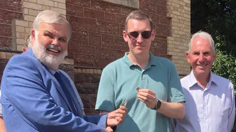 A group of three men stood in front of a monastery, all with smiles on their faces. The man on the left has grey hair and a grey beard, and is wearing a royal blue coloured suit and holding a key. The man in the middle is holding a pound, and is wearing a light green polo top and sunglasses. He has brown hair. The man on the right has grey hair and is wearing a purple toned button up shirt.