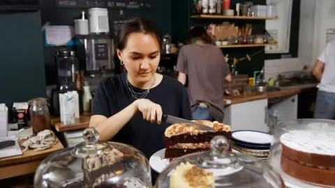 Getty Images A young woman wearing a black T-shirt stands behind a counter of cakes in a cafe, cutting a large slice of cake
