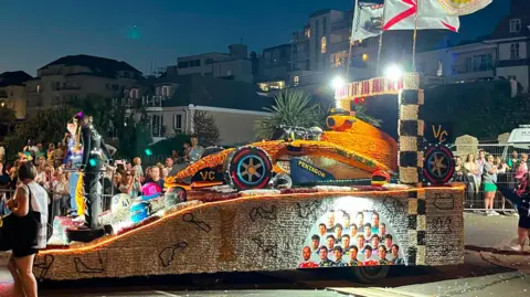 BBC An illuminated Jersey Battle of Flowers parade float shaped like a Formula One racing car travels along a street at night. Spectators line both sides of the road behind barriers, while flags fly above the float and buildings are visible in the background.