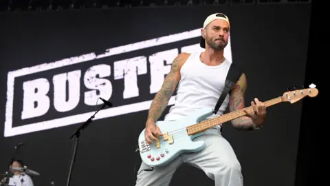 Getty Images Busted's Matt Willis is on stage playing a very light blue bass guitar. He wears white jeans, a white vest and a white backwards cap. He stands behind a microphone, in front of a screen that reads "Busted" in white letters.