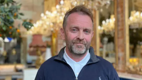 Eamon McCusker standing in front of a row of mirrors and chandeliers in the Chubby Cherub restaurant He is wearing a navy half-zip fleece over a white polo shirt. 