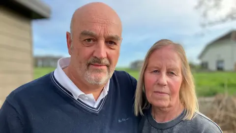 Mark and Sandra Thompson are standing outside in front of a static caravan. Mark is wearing a dark blue sweater over a white collared shirt, and Sandra is wearing a gray sweatshirt. The background shows green grass, another building, and a partly cloudy sky.