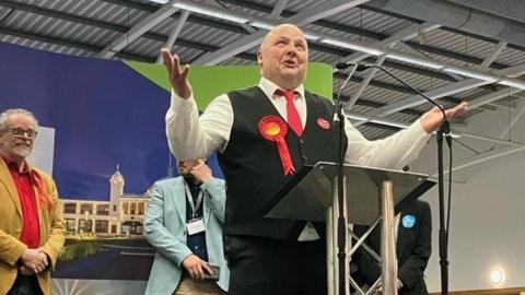 Andy Holdsworth, wearing a red rosette, giving a speech 