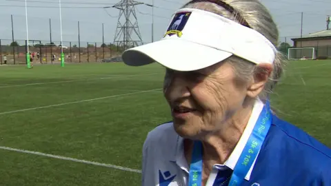 Wendy Watson, wearing a white visor cap and her grey hair pulled back. She has a 5k & 10k fun run lanyard on and a blue and white t shirt. She is standing on a rugby football pitch.