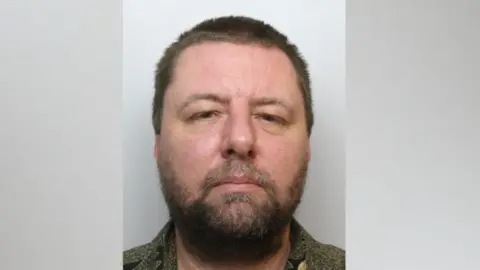 Mugshot of a man in a beard and looking seriously at the camera.