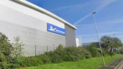 A large silver-coloured warehouse building sits behind a security fence with the Michelin logo on the side of it. In the foreground there is a grass verge and some bushes as well as  lampposts at the side of a pavement.