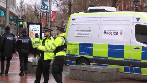 Police officers stood outside vans in Reading town centre with a van that says "facial recognition currently in use" and Thames Valley Police's logo next to it.