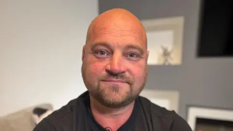 BBC Offshore worker Phil Perry, who has a bald head and is wearing a black T-shirt, is smiling at the  camera. He has tattoos on arms.