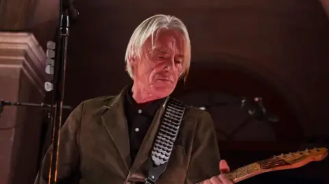 A man with long grey hair, a brown jacket and a brown guitar performs on stage. He looks towards the head of his guitar while a black microphone stand can be seen in front of him.