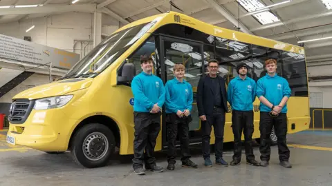 Two young men wearing a blue sweatshirt and black trousers stand to the left of Mayor of Greater Manchester, Andy Burnham, and two other young men stand to his right, all are standing in front of a yellow bus