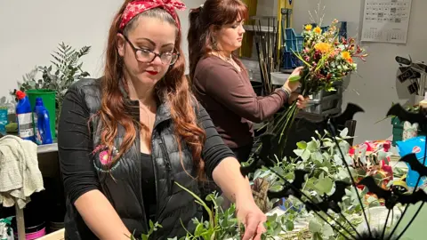 Two young women, both with dark auburn hair are working inside the florist. One stands closer to the camera, she is focusing on an arrangement and wears a red and white bandana in her hair with large black framed glasses, a long sleeved black top and black gilet over the top. Just behind her, the other woman wears her hair in a ponytail and is holding a bouquet of flowers with a mixture of orange and red buds and evergreens.