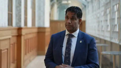 Un hombre con traje azul marino y corbata está de pie en un pasillo, mirando a la cámara. Tiene las manos entrelazadas. Al fondo, tras él, se ven paneles de vidrio y madera.