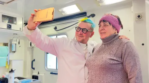 Serhii Baksheiev A doctor smiles as he takes a selfie with a woman patient inside a mobile medical unit. His hair is dyed the blue and yellow of the Ukrainian flag, and medical paraphernalia is in the background.