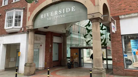 The front of a shopping centre - there are two concrete pillars, a glass frontage and a sign that says 'Riverside Shopping Centre'. 