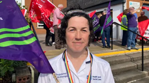 Maeve O'Neill has curly black hair and is wearing white medical scrubs. She is holding a large purple and green flag and a number of people on a picket line are visible behind her. She is standing near steps outside Altnagelvin Hospital in Derry. 