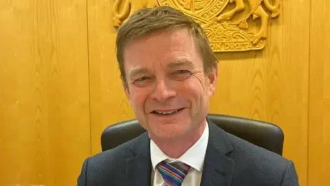 Judge Patrick Perusko sitting on a chair in court. He is smiling at the camera and is wearing a dark suit, white shirt and multi-coloured stripped tie. A coat of arms is behind him on a wood-effect wall. 