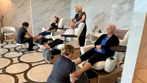 Three women sit on chairs in a spa with an ornate marble floor and have their feet massaged by the staff