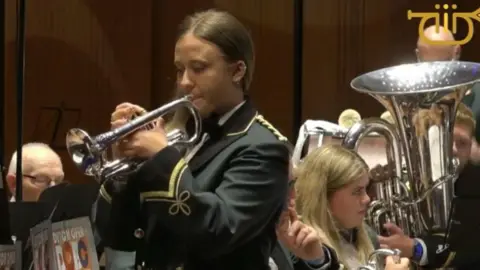 A young girl is pictured from the waist up. She is wearing a white shirt and green blazer with gold threaded embellishments. She has long brunette hair and is holding a silver cornet to her mouth. She is standing in front of a brass band - a large tuba and a few other band members can be seen behind her. 