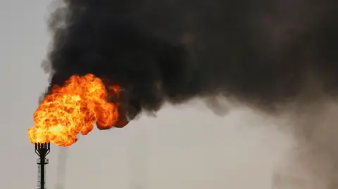 Reuters Blooms of bright orange fire leading to thick black smoke burst out from a metallic pipe in a grey sky.