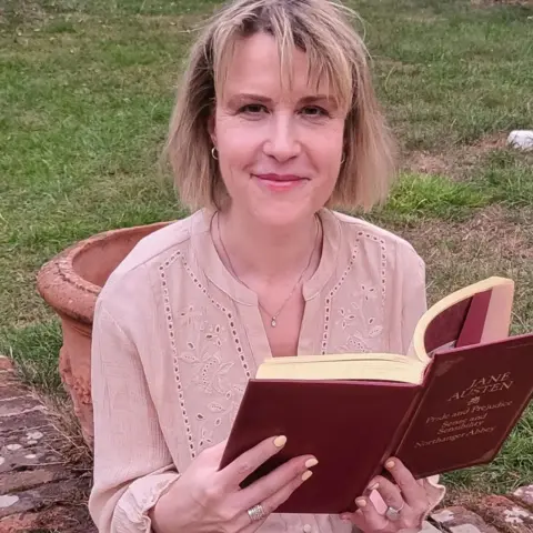 Joanne Wenman Joanne wears a light pink blouse and is sitting in a garden, holding a red hardback copy of an Austen book compilation.