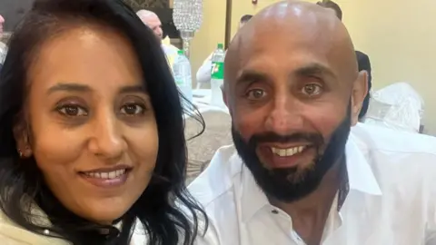 Samina Zaman Samina Zaman with long black hair and a striped black and white top. She is posing with a man who has a bald head and thick black beard and moustache. They are both smiling.