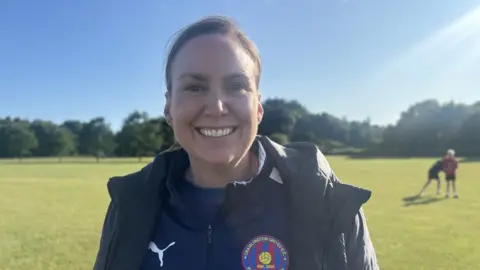 A women who is smiling with dark brunette hair, wearing a black puma jacket with the crest of cramlington united on the left hand side of the jacket. Her background is a green field with trees in the distance.
