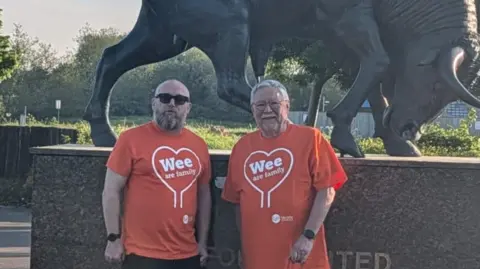 Neil McManus Neil McManus and another man are wearing orange t-shirt and are standing in front of the bronze Ox statue outside Oxford United's Kassam Stadium.