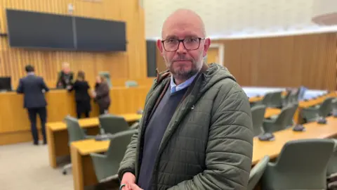 Bald man with glasses, green puffer jacket stands in empty council house chamber