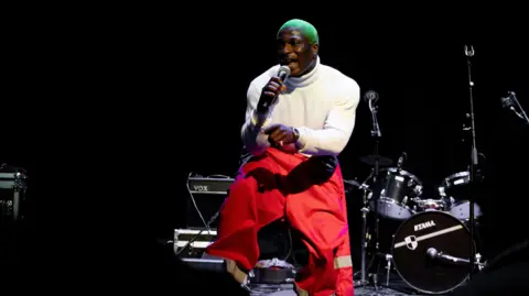 Jack Oliver Sambee performing at an event on a low lit stage, wearing a white roll neck, red trousers and dyed green hair.