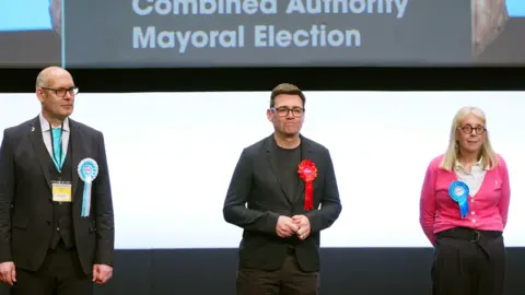 PA Media Andy Burnham (centre) is re-elected as Greater Manchester Mayor in 2024 as Reform candidate Dan Barker and Conservative candidate Laura Evans look on. Burnham wears a black jacket and trousers with a black t-shirt.