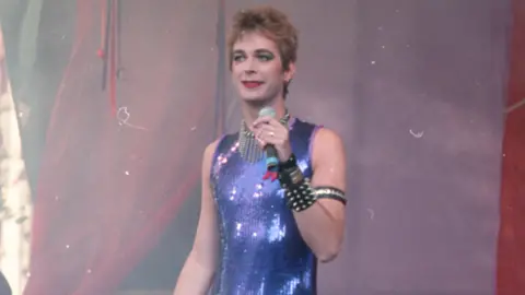 Stuart Rhoades Julian Clary, who is wearing a purple top and jewellery on stage. He his holding a microphone up to his mouth. 