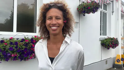 A woman with brown curly hair. She has a white top on. She is smiling at the camera. Behind her is a white building with flowers under the windows.