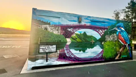 Telford and Wrekin Council A graffitied hut of an iron bridge with grassy banks over a river. There is also a painting of a large kingfisher. The hut is on a car park with the sun setting behind a hill in the background