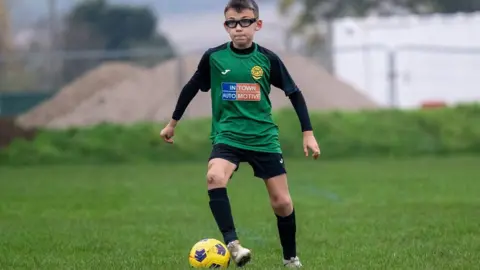 Fewster family Ezra wearing his glasses and a green and black football strip. He is kicking a yellow and blue ball across the grass.  Metal fencing is visible behind him.