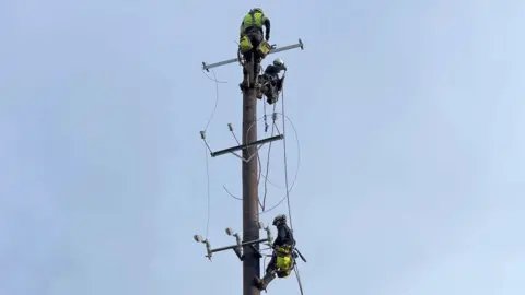 PA Media A wide shot of an electricity pole against a grey-blue sky, three workers in high-viz and helmets are working on it