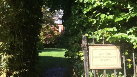 A path between an overhanging hedge leading to a lawn with a red brick building in the background. At the entrance to the path there is a sign which reads: "Hill End Commemorative Garden"