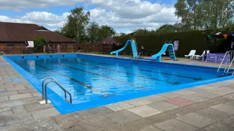 A view of the pool.  It has a bright blue surround and the water is crystal clear.  Two slides go into the pool on the side and there are steps at each corner.