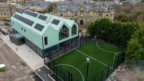 A large two-storey building with a mint-coloured roof and large windows sits next to a sports court with an artificial grass surface. Buildings can be seen on the road behind.