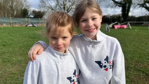 Two young girls stand smiling in a dog agility field. They both have light brown hair tied back in a ponytail, and wear grey zip-through jumpers with a logo of a dog on the chest. In the background, tunnels, jumps and other obstacles can be seen. 