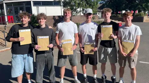 Phil Harrison/BBC Six male students stand outdoors in a school playground holding large brown envelopes on a sunny day.