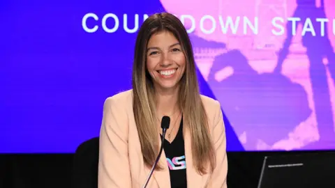 NASA A woman wearing a cream coloured blazer, smiling. There is a black microphone in front of her, with an LED background of blue and purple, with text in white that reads "countdown".