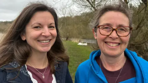 Two women are pictured on a grassy patch of land in front of two swans. The woman on the left has long brown hair, a burgundy top and is smiling. The woman on the right is wearing a bright hooded top and has a pair of tortoiseshell glasses. 