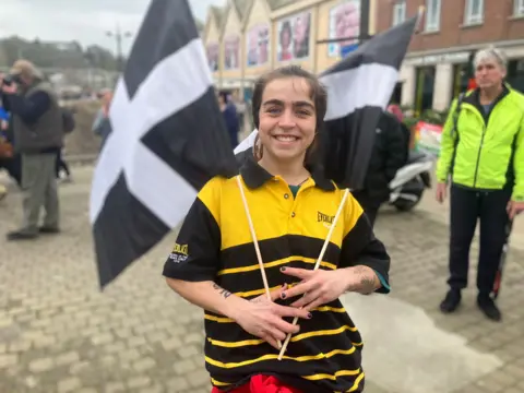 A picture of a woman holding Cornwall flags and wearing a black and yellow polo