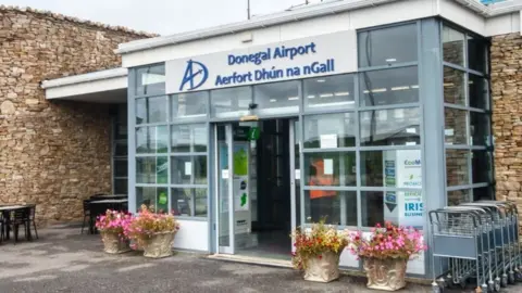 The front of Donegal airport, it is a small building with a glass entrance porch and stone walls on either side. The sign on the porch reads "Donegal Airport" in blue font on a grey background, with the Irish translation underneath. 