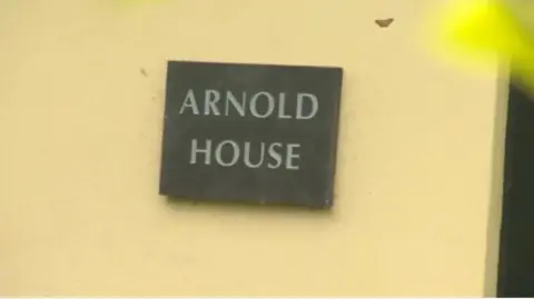 A yellow wall with a black slate sign hanging that reads 'Arnold House'