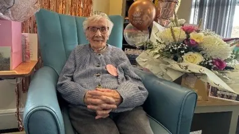 Bloomfield Court care home An elderly woman sat in a big blue arm chair in a blue cardigan smiling, with gifts, flowers and rose gold, pink and clear balloons. 