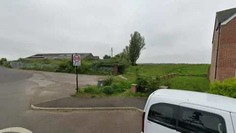 Street view of a green field with a mast in the distance. In the foreground on the left, there is an industrial building, and to the right, there is the edge of a red-bricked two-storey house.