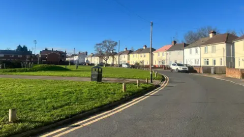 BBC A road bend next to a children's park with a footpath running between it. On the other side of the road there's a row of attached houses, a couple of them have white cars parked in front of them. It's a sunny bright day with a clear blue sky. 