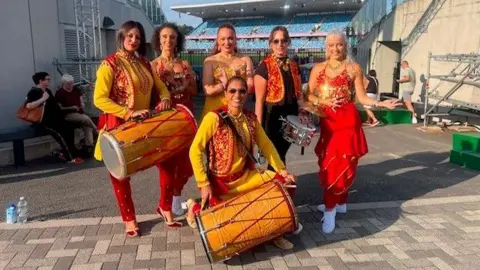 Eternal Taal Five women standing up, while one kneels in front of them. They are all dressed in red and yellow outfits with vines and leaves on the tops. The woman on the left is holding a large dhol drum. The woman ont he floor is also holding a dhol drum