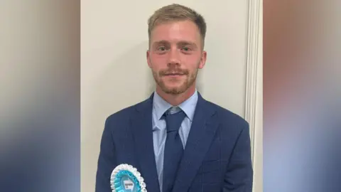 Reform UK councillor for Harraby South ward Josh Kirkwood. He is wearing a blue suit and tie and pale blue and white rosette.